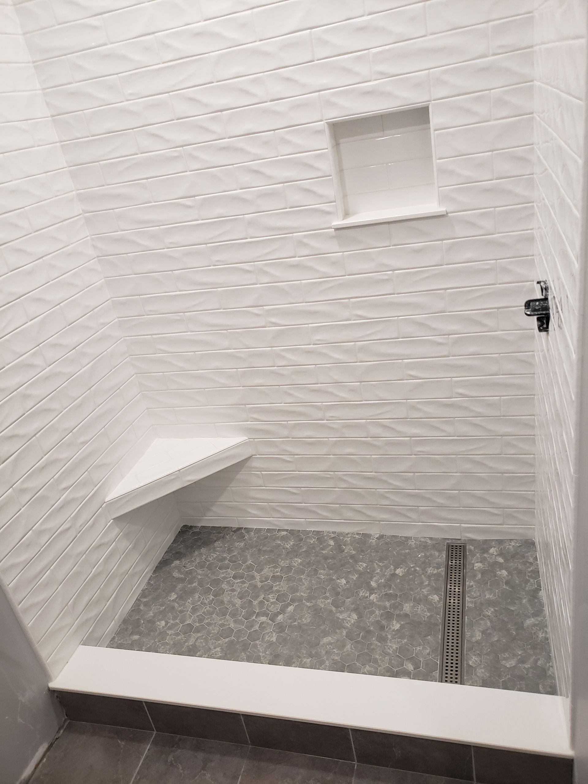A walk-in shower with textured white tile walls, a gray stone floor, a corner seat, a small inset shelf, and a linear drain.