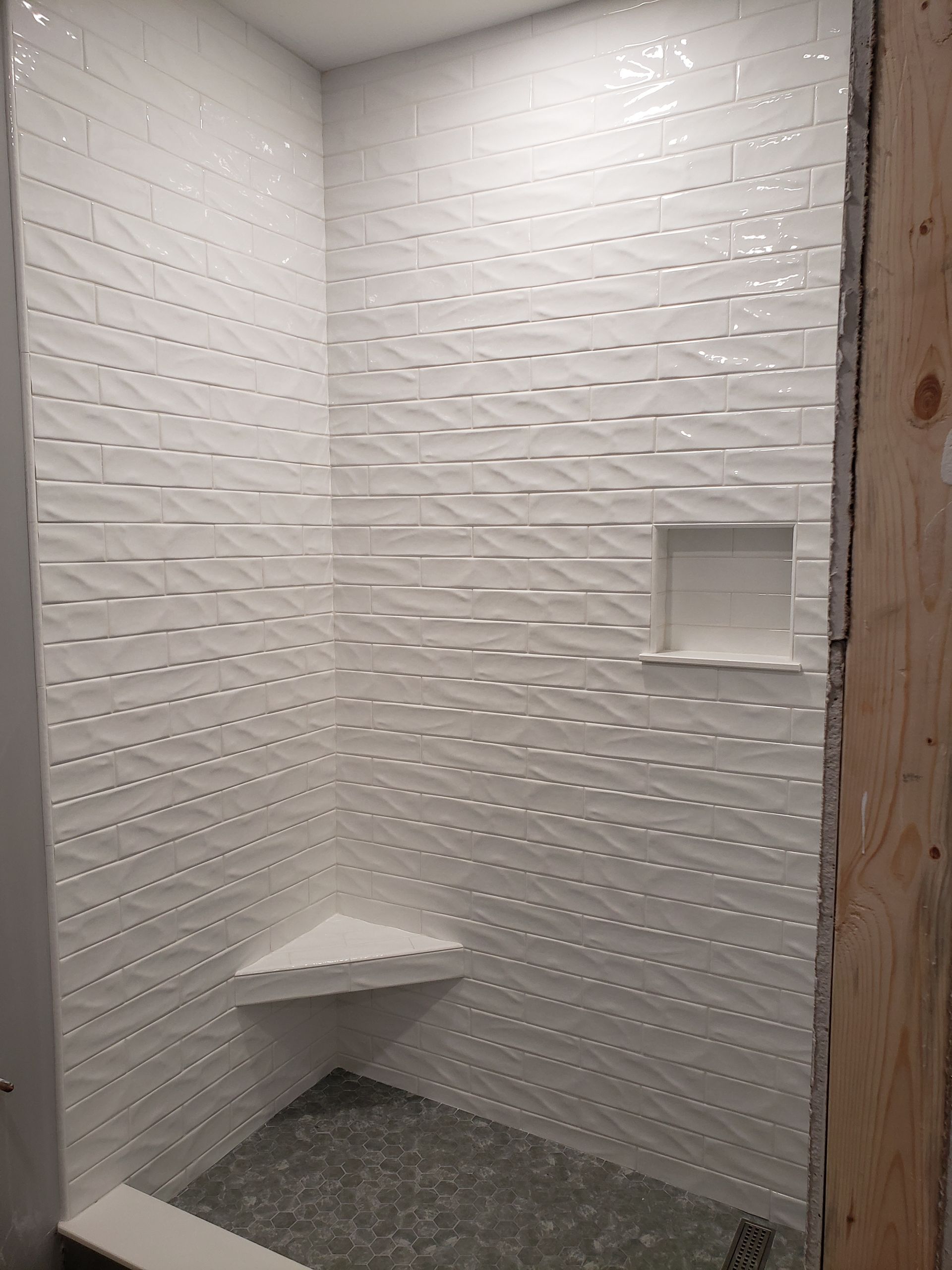 A tiled shower stall with white wavy-textured wall tiles, a corner seat, and a small built-in niche above a dark floor.