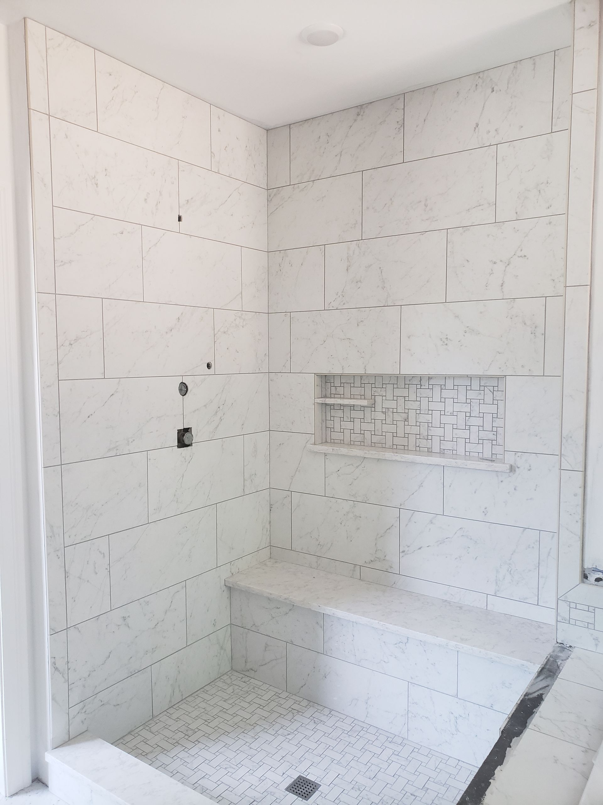 A tiled shower stall under construction with white marble-patterned wall tiles, a built-in bench, and a mosaic floor.