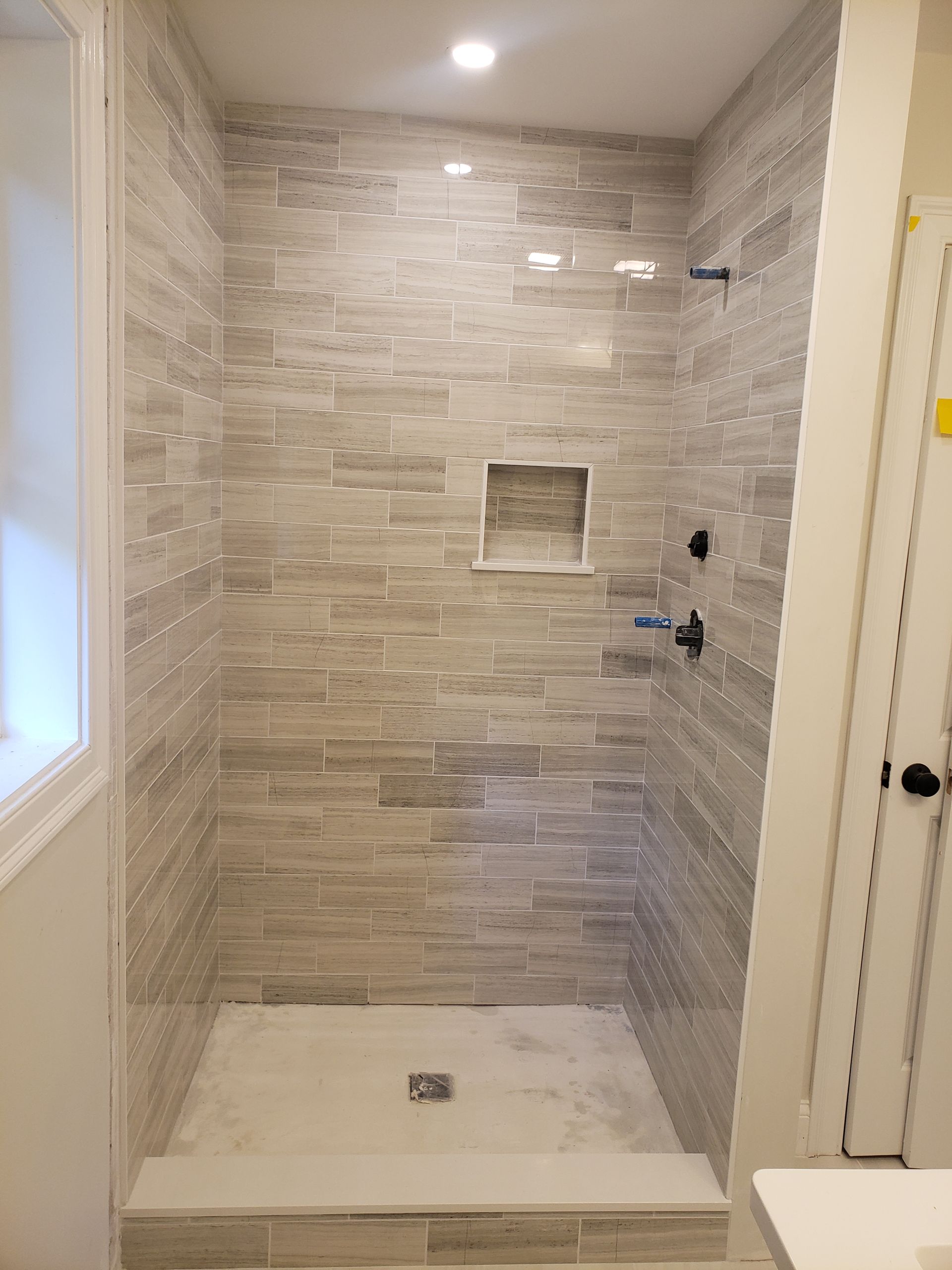 A walk-in shower with light grey, rectangular tiled walls, a square inset shelf, and a white tiled floor.