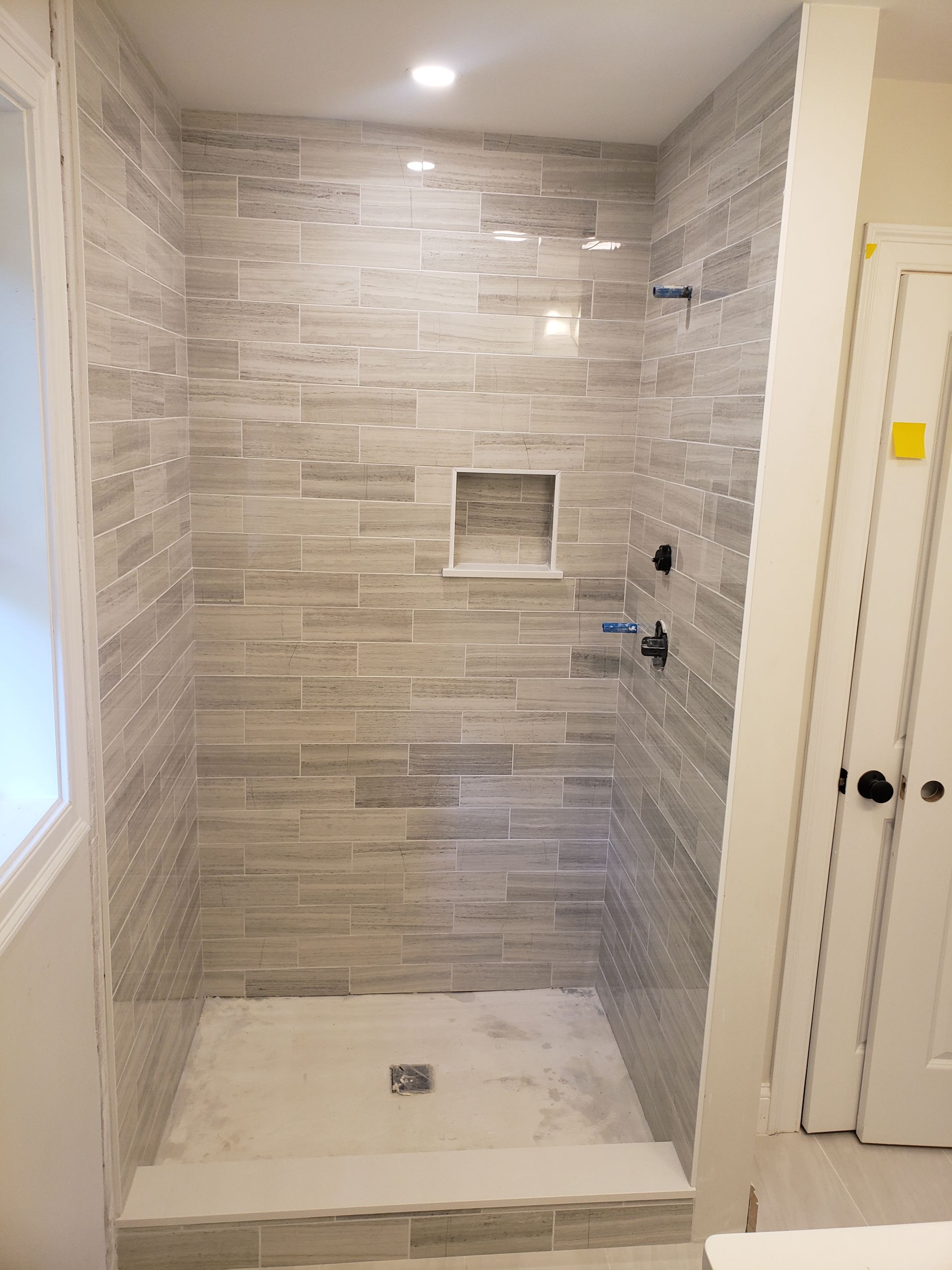 A tiled shower stall with grey, textured horizontal wall tiles, a built-in recessed shelf, and a light-colored shower pan.