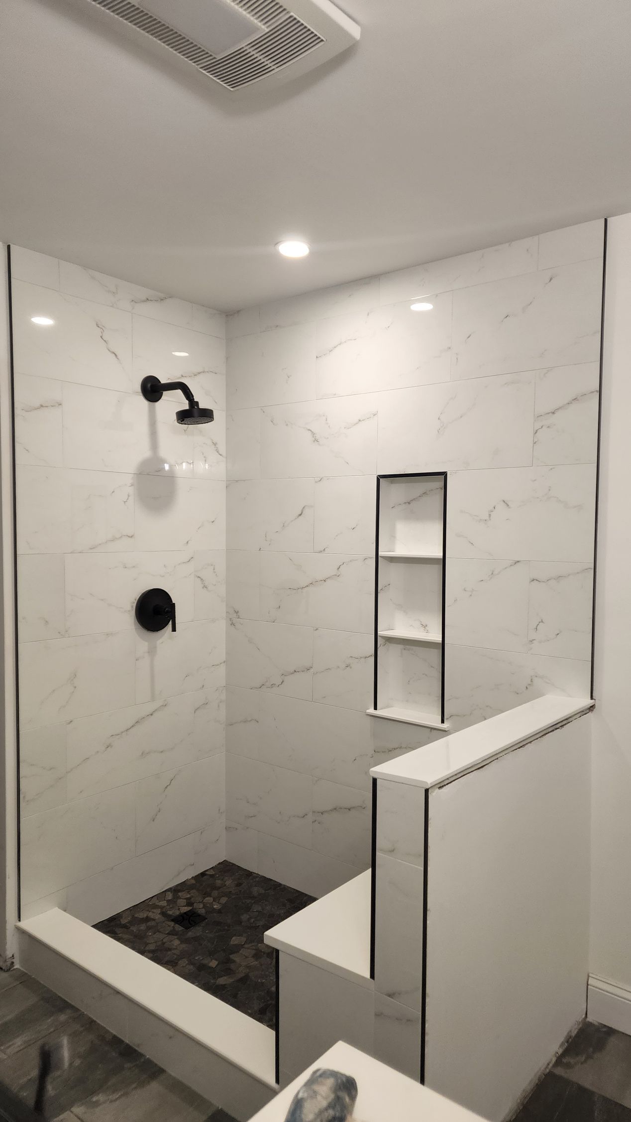 A modern walk-in shower with white marble-patterned walls, a dark stone floor, a built-in bench, and black fixtures.