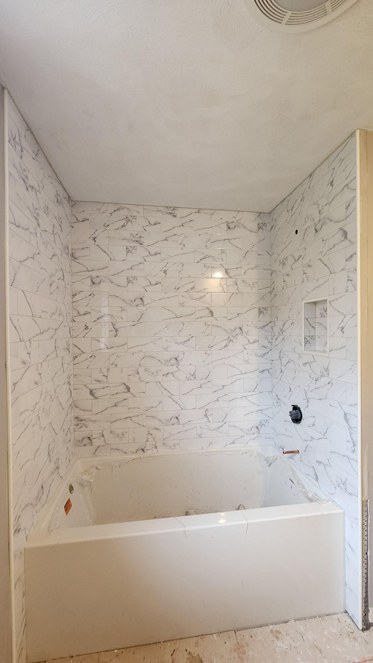A bathroom shower-tub surround featuring white panels with dark grey marble-look veining, a niche, and black hardware.