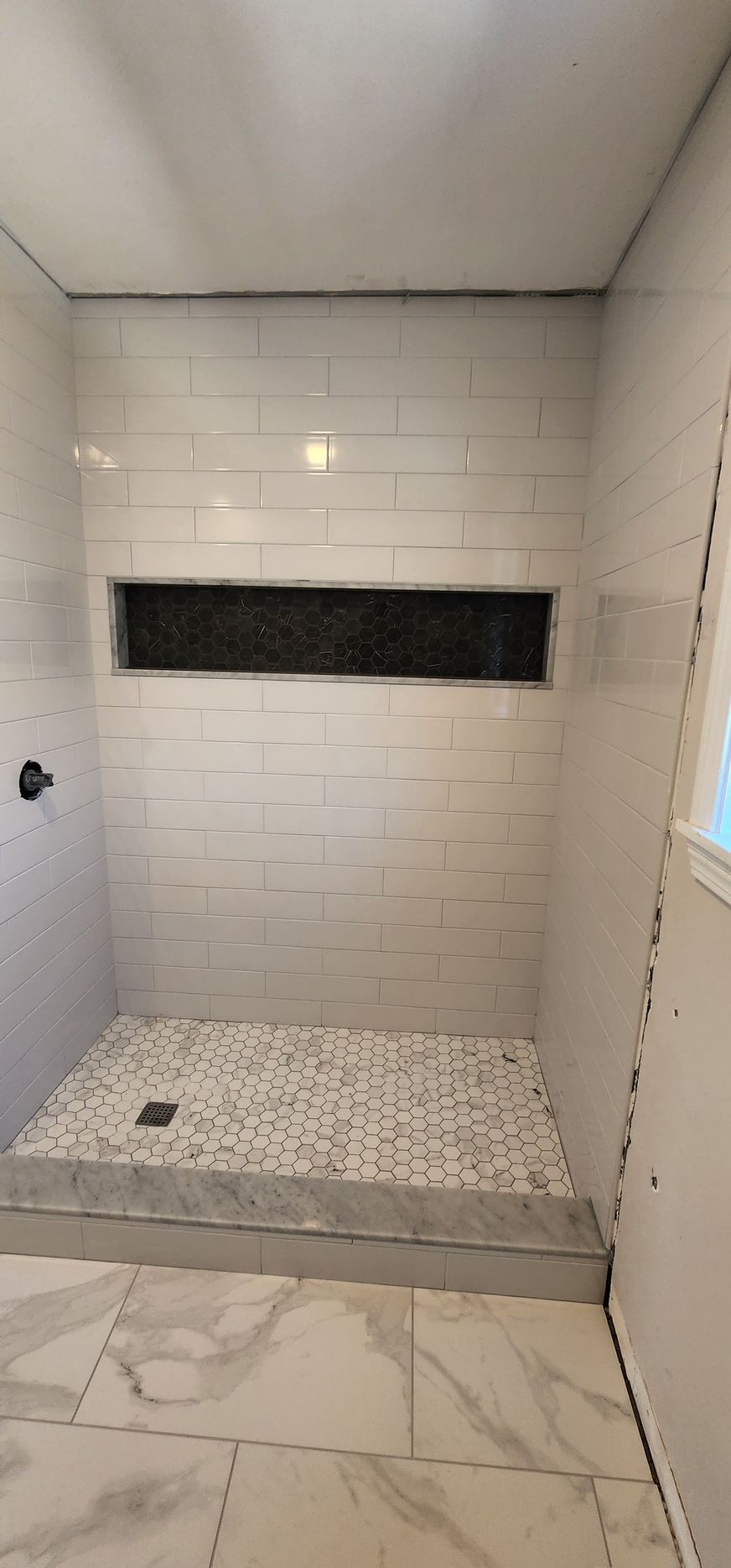 A shower with a white subway tiled back wall featuring a dark niche, a decorative tiled floor, and a marble-look step.