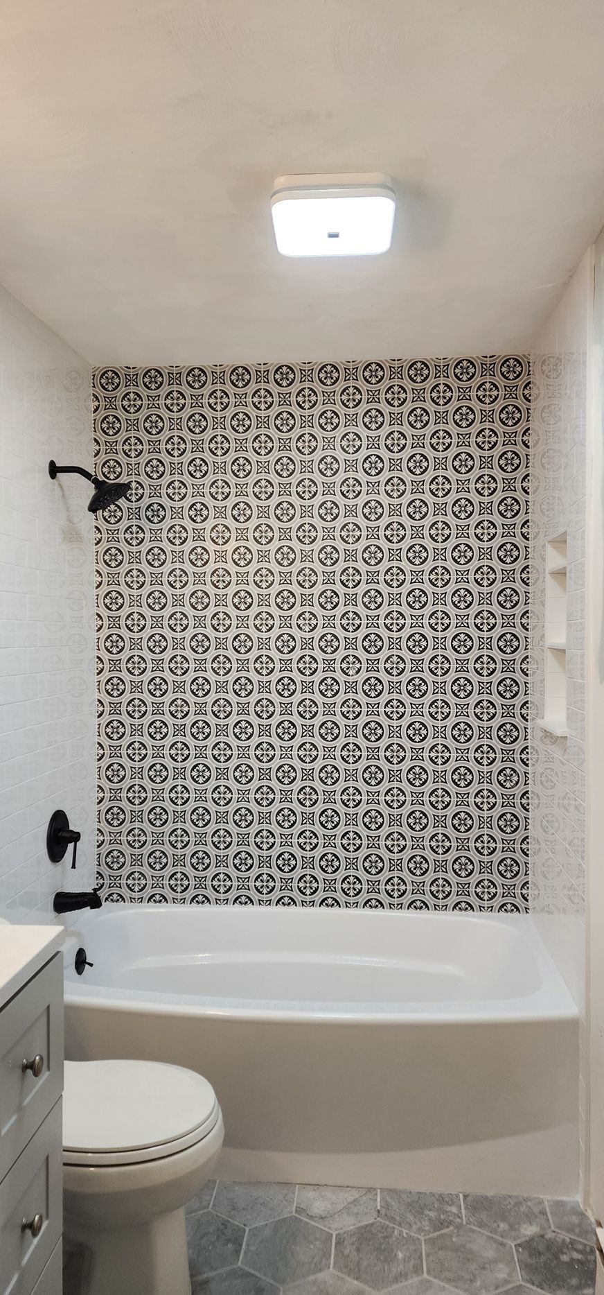 A bathroom featuring a white bathtub with a black-and-white patterned tile accent wall, matte black fixtures, and grey floor.