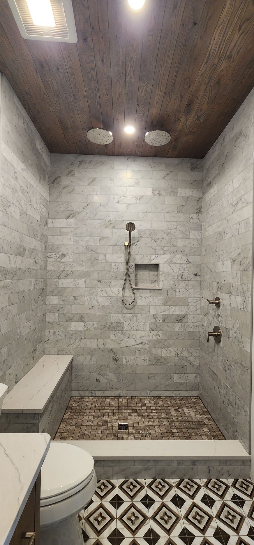 A walk-in shower with light grey tile walls, a dark wood ceiling, a pebble floor, a built-in bench, and geometric tiles.