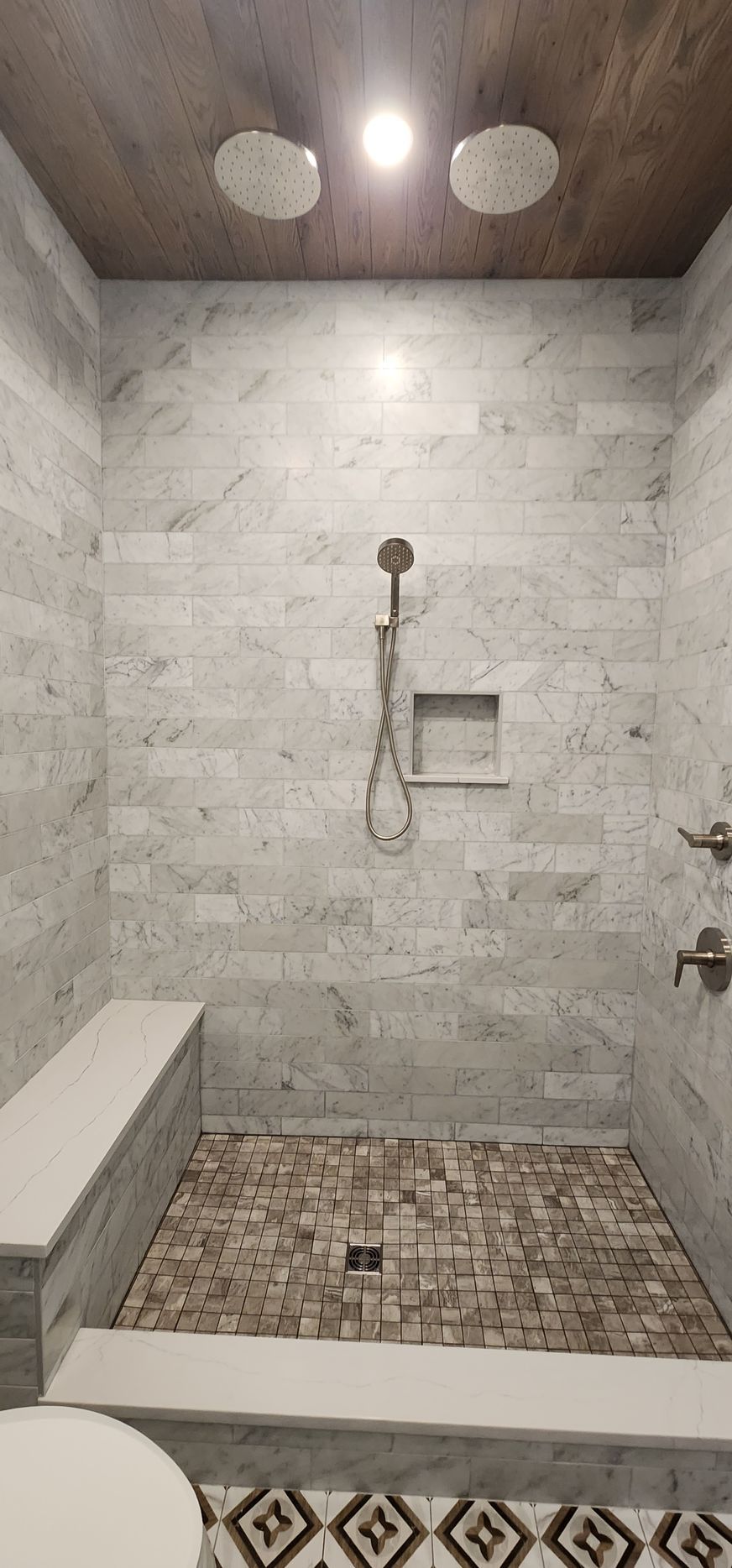 A modern walk-in shower with light gray marble walls, a wood-look ceiling, dual showerheads, and a stone-tiled floor.