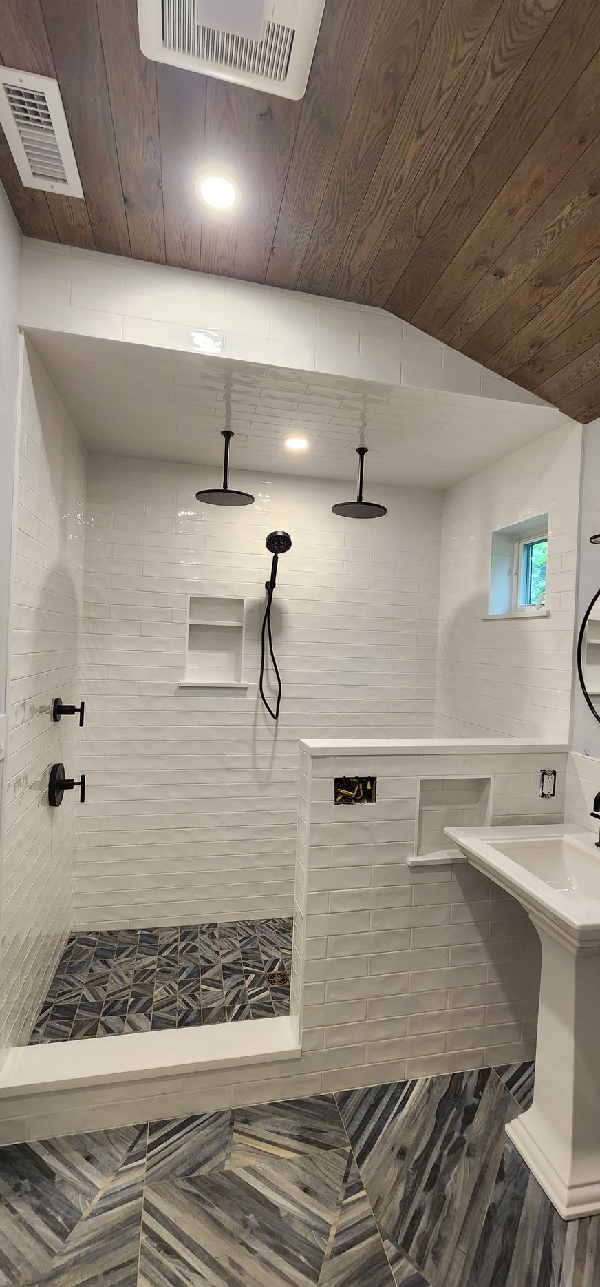 Modern bathroom featuring a walk-in shower with white tile walls, dark stone floors, and black fixtures under a wood ceiling.