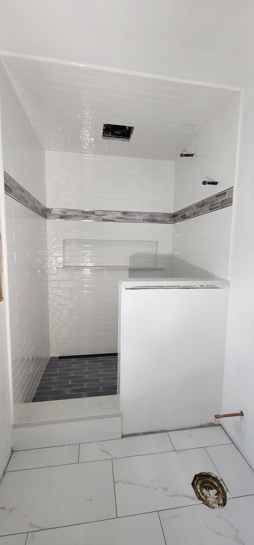 A walk-in shower with white tiled walls, a decorative border, and a built-in bench, unfinished in a bathroom with floor tiles.