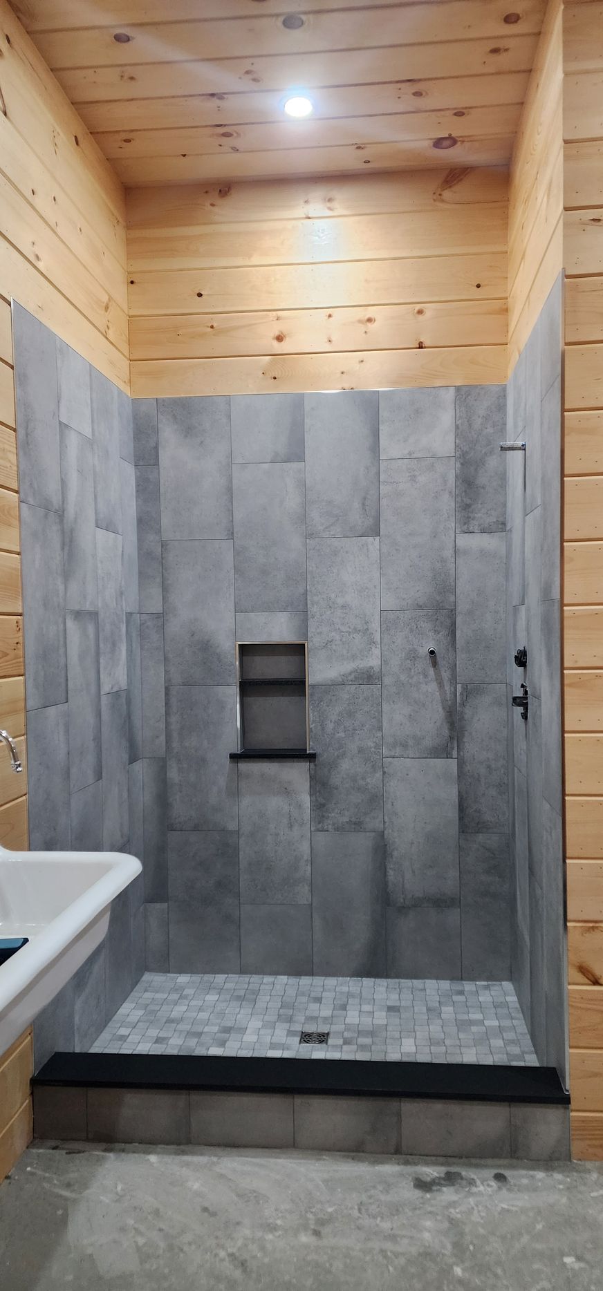 A shower with grey vertical tile walls, a dark recessed shelf, and a dark curb against natural wood-paneled walls.