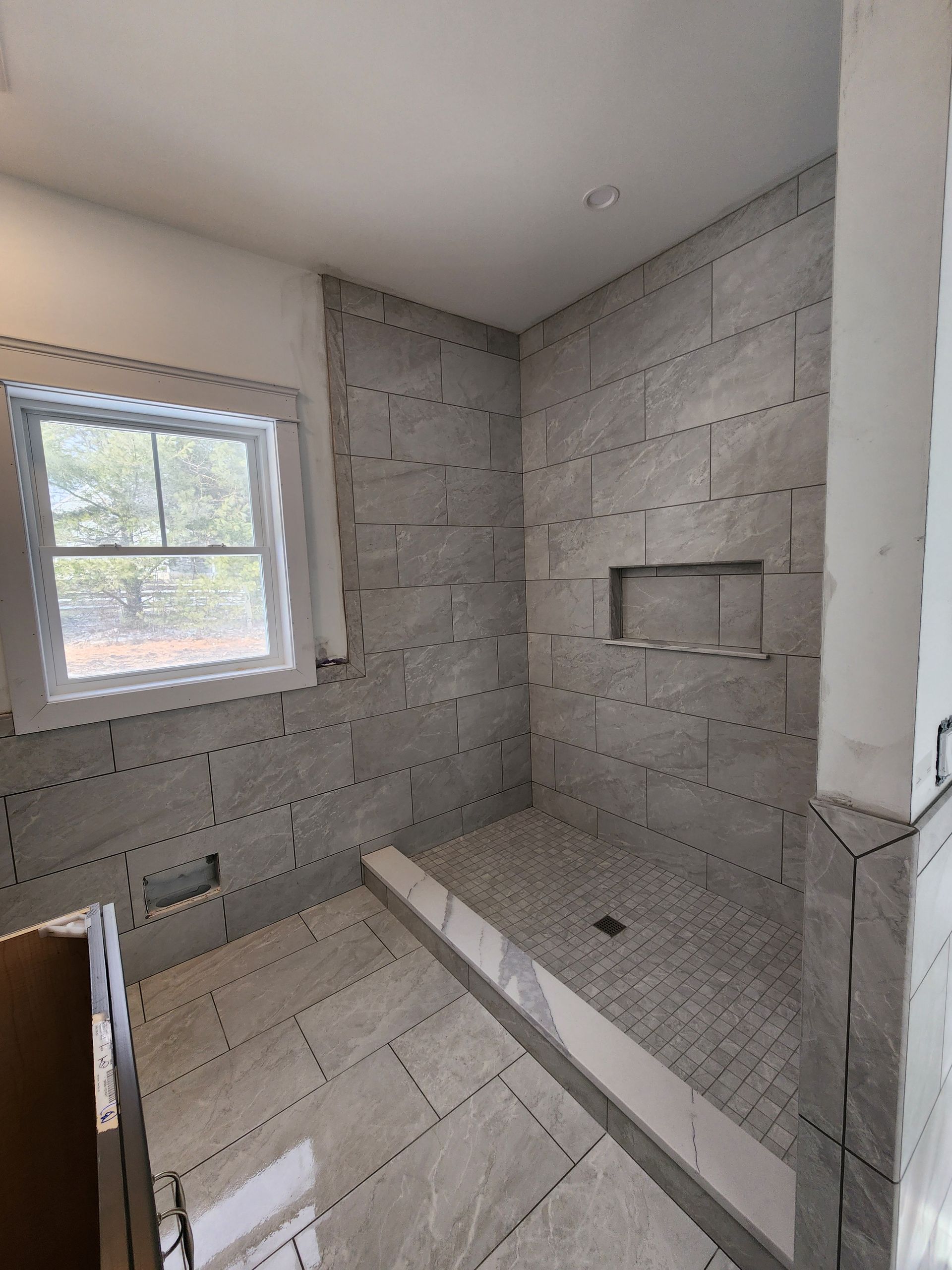 A partially tiled walk-in shower with a window, niche, and light-gray stone flooring in a room under construction.