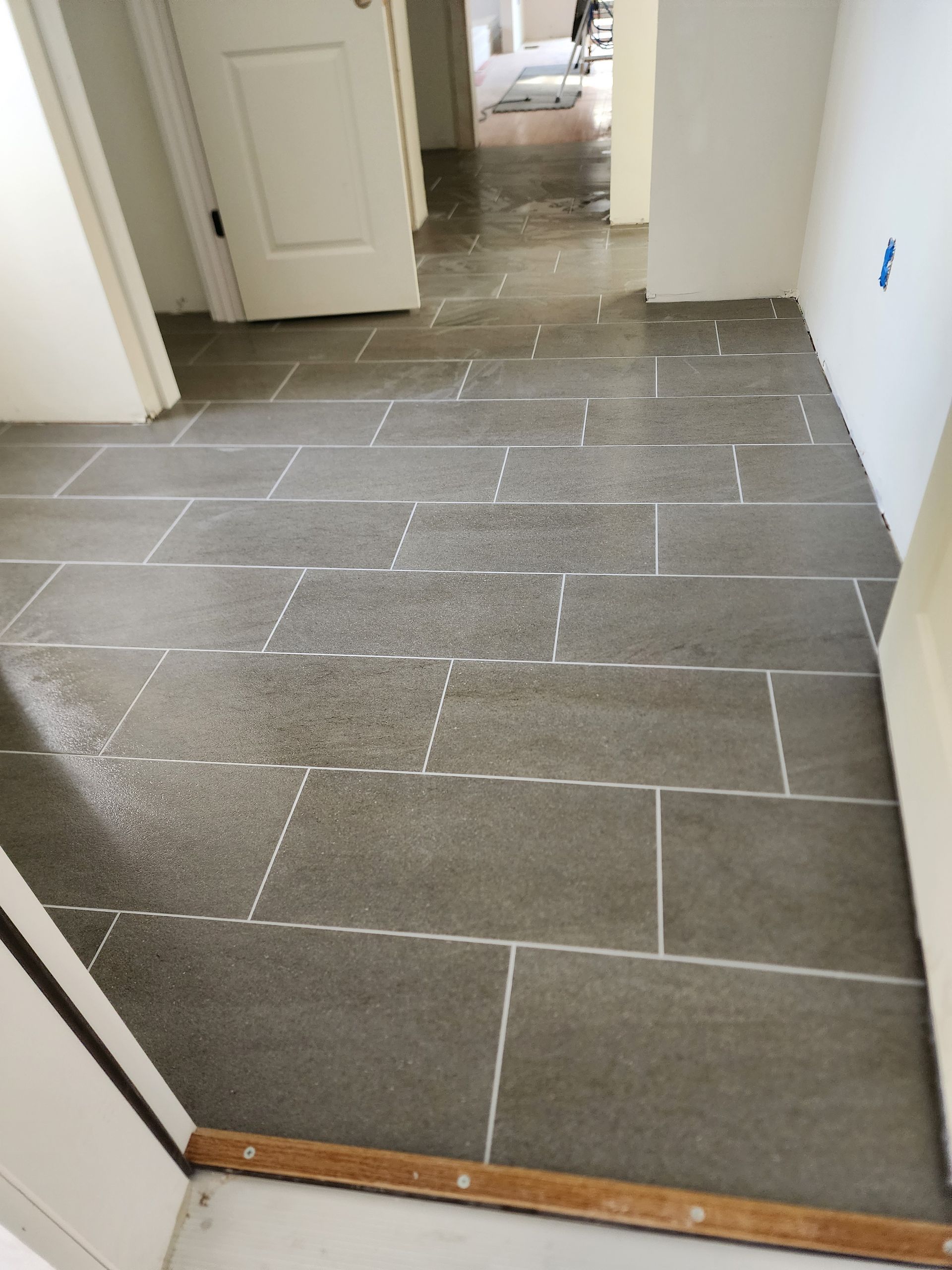 A hallway with grey rectangular floor tiles installed in a running bond pattern, leading toward an open doorway.