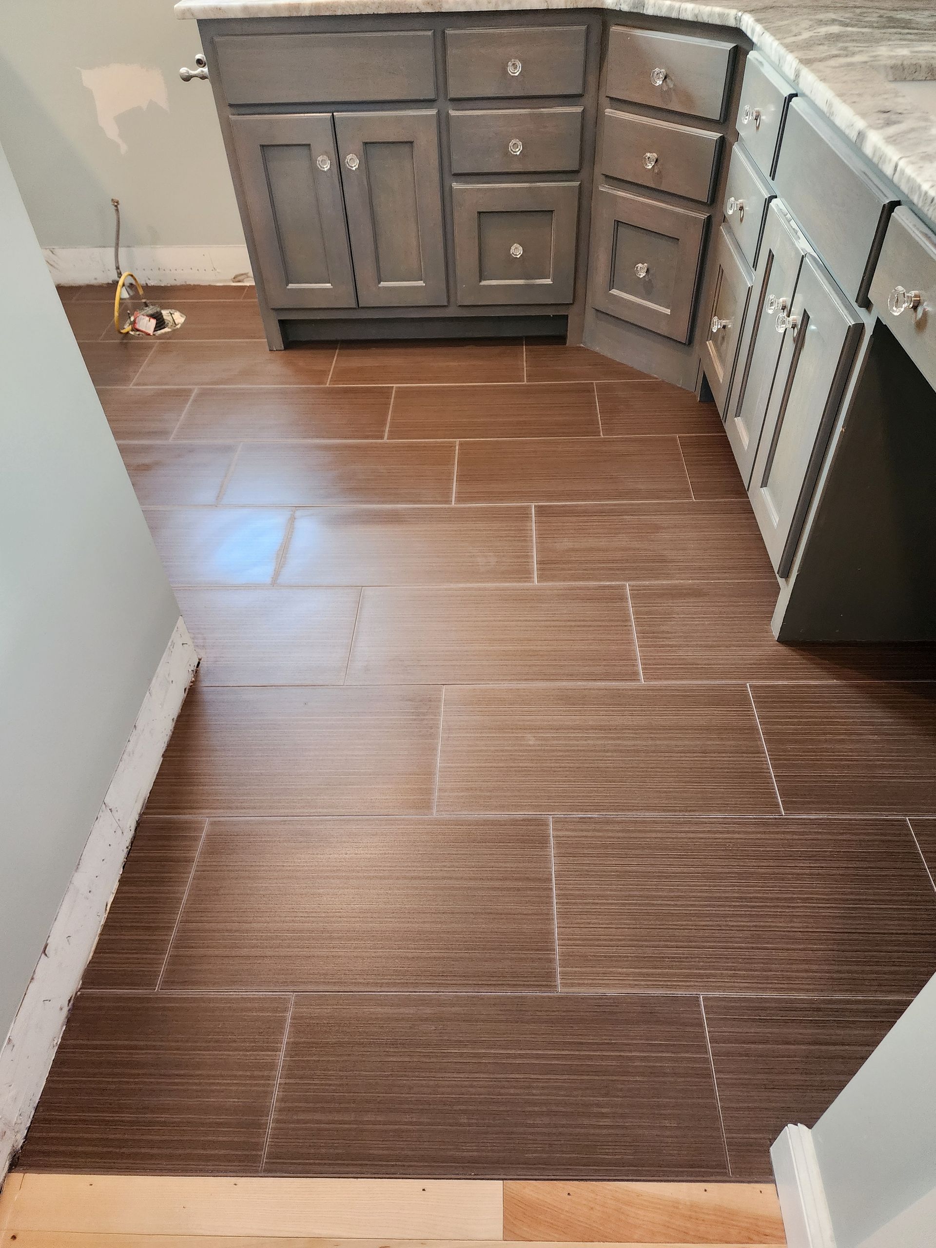 A partial view of a kitchen featuring brown horizontal-patterned floor tiles installed in front of grey wooden cabinets.
