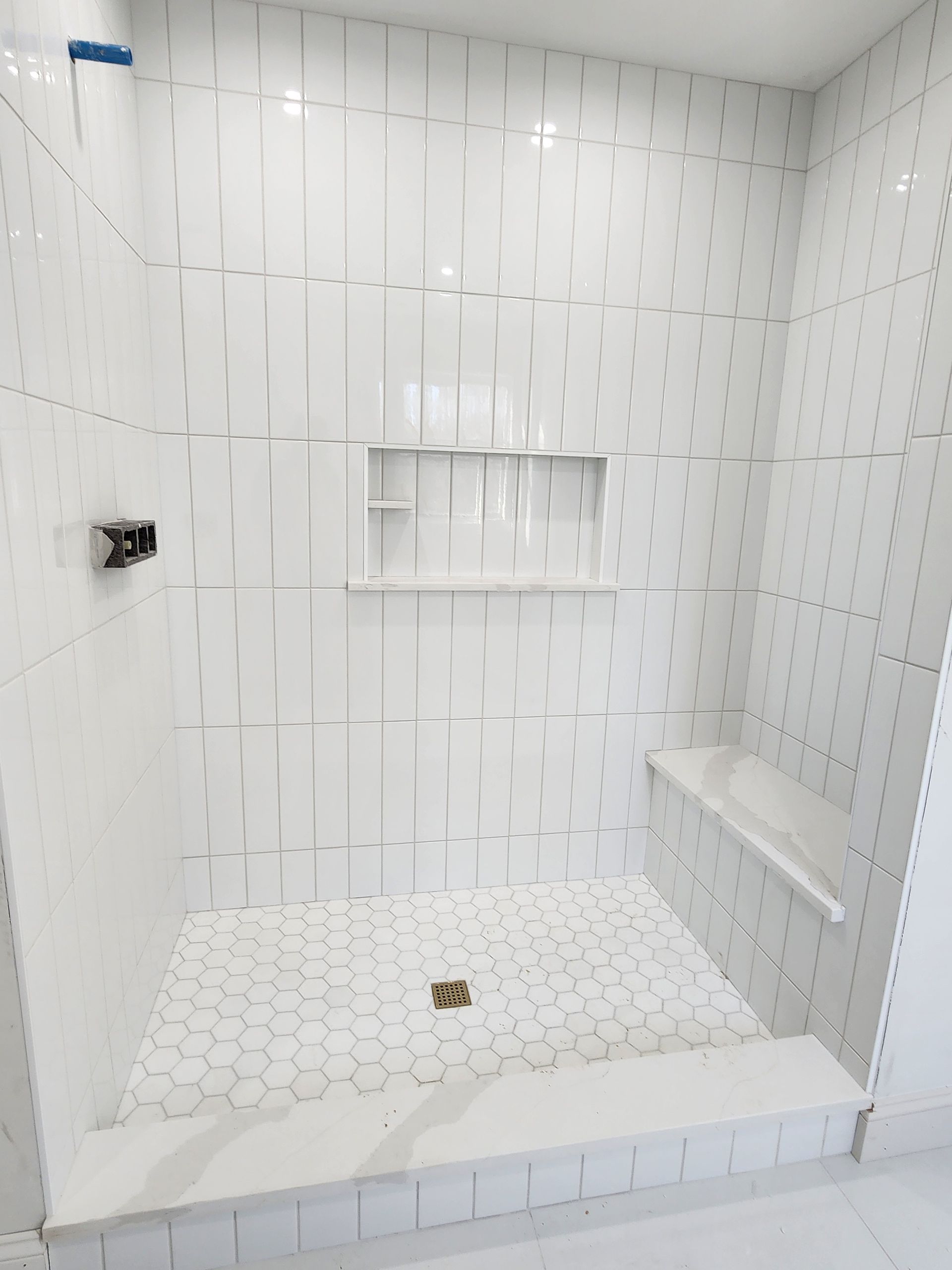 A tiled shower interior with white rectangular wall tiles, a built-in recessed shelf, a bench, and a hexagonal floor pattern.