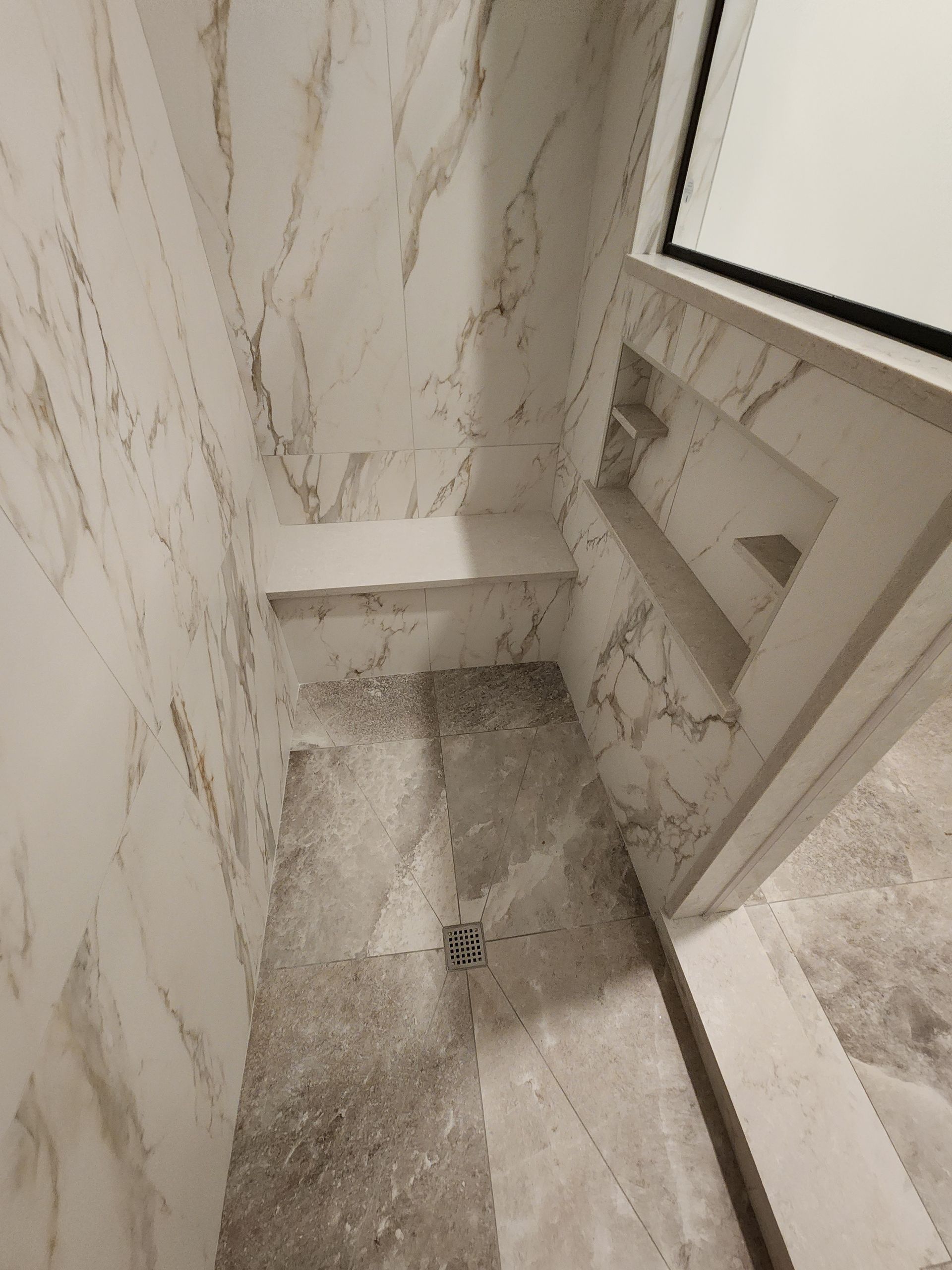 A modern shower with white marble walls, a built-in bench, a recessed shelf, and light stone tile flooring.