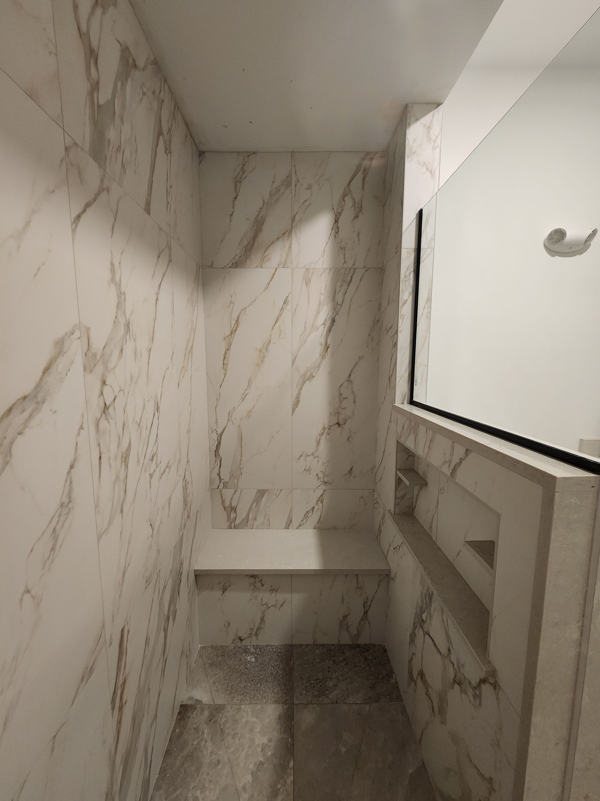 A marble-tiled walk-in shower with a built-in bench and a recessed niche shelf under a mirror.