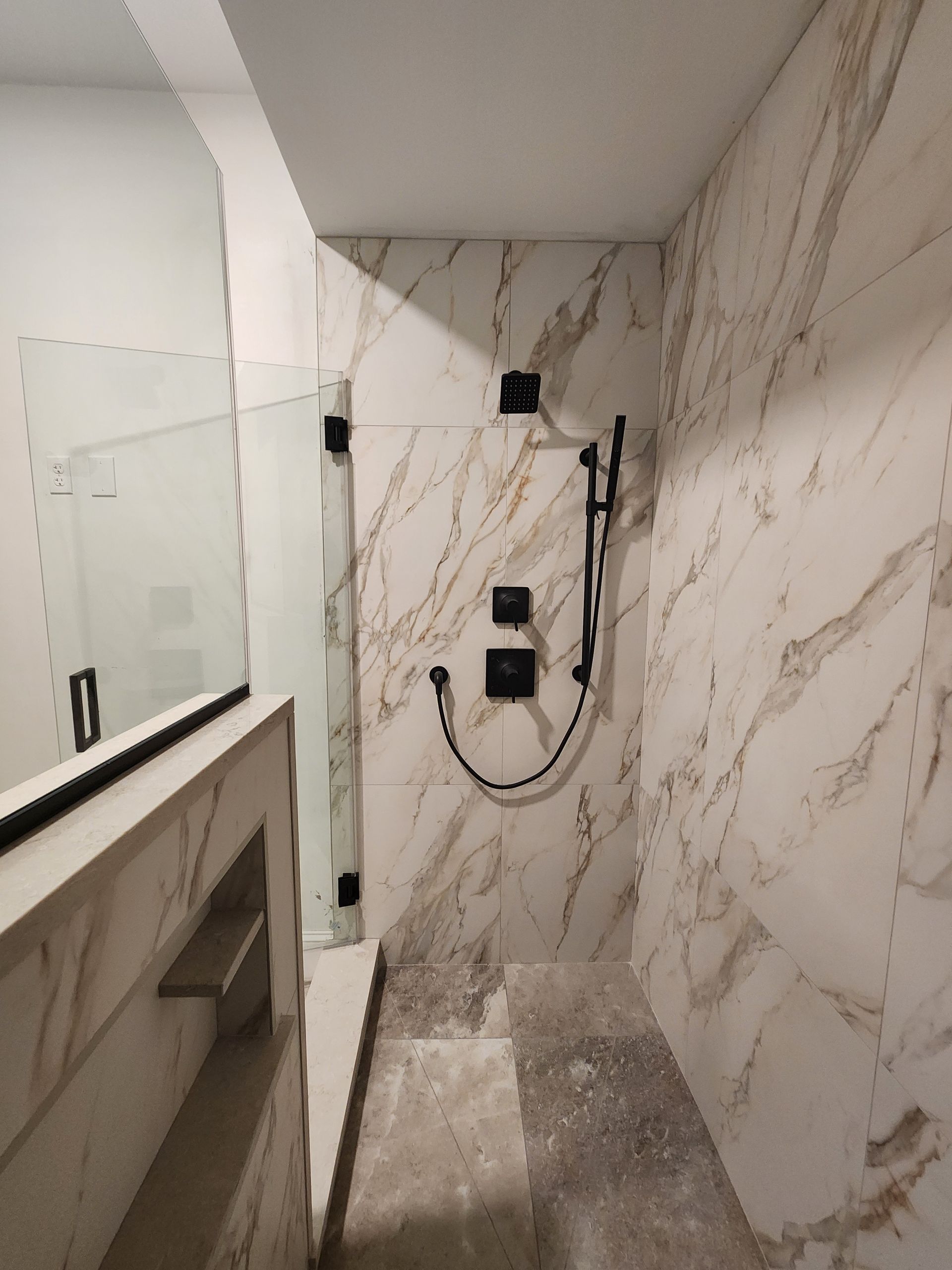 A shower stall with white and brown marble tiles, matte black fixtures, a handheld sprayer, and a glass partition.