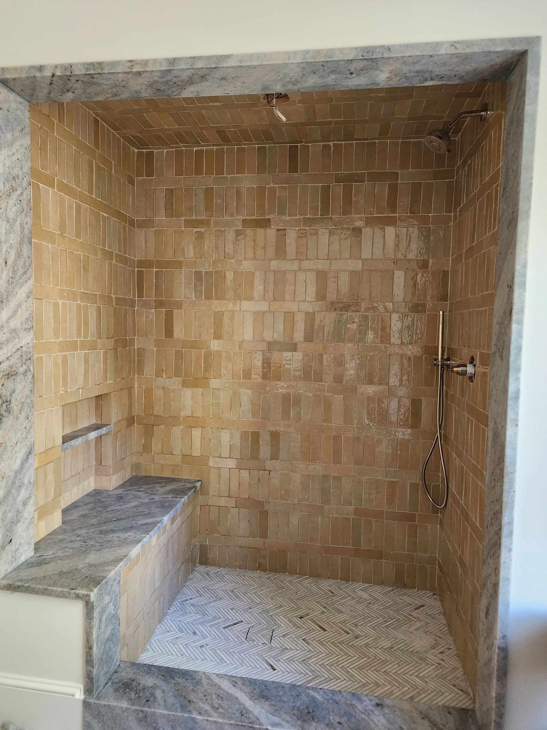 A walk-in shower with tan rectangular brick-style wall tiles, a light-colored pebble floor, and grey stone trim and bench.