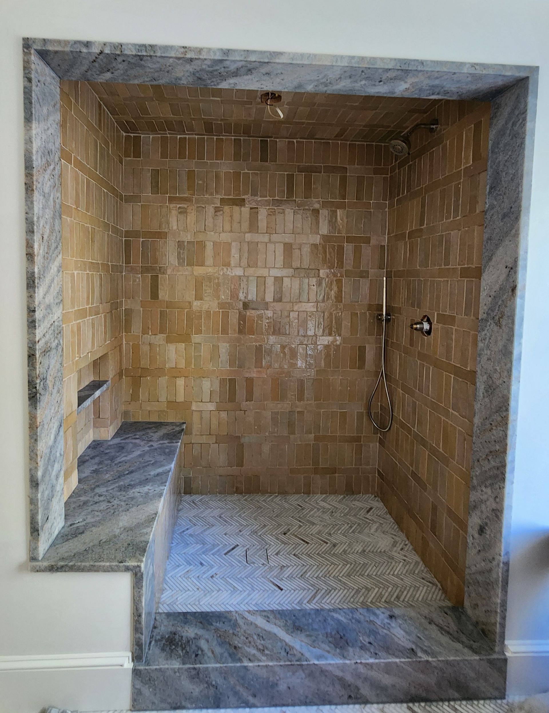 A walk-in shower with tan rectangular wall tiles, a mosaic floor, and grey-veined stone trim, seat, and threshold.