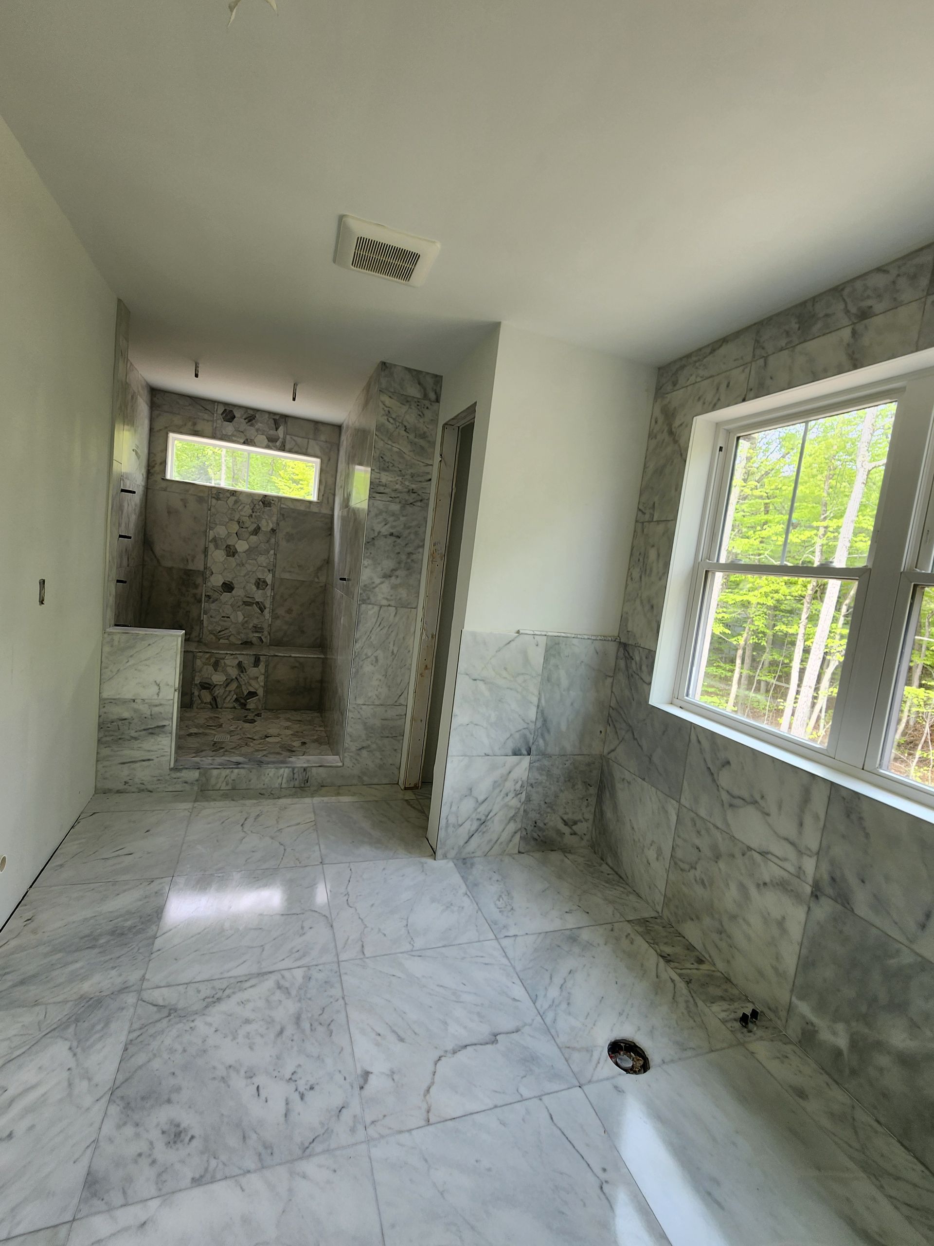 An unfinished bathroom featuring marble tiled walls and floors, a glass-less walk-in shower, and a large window.