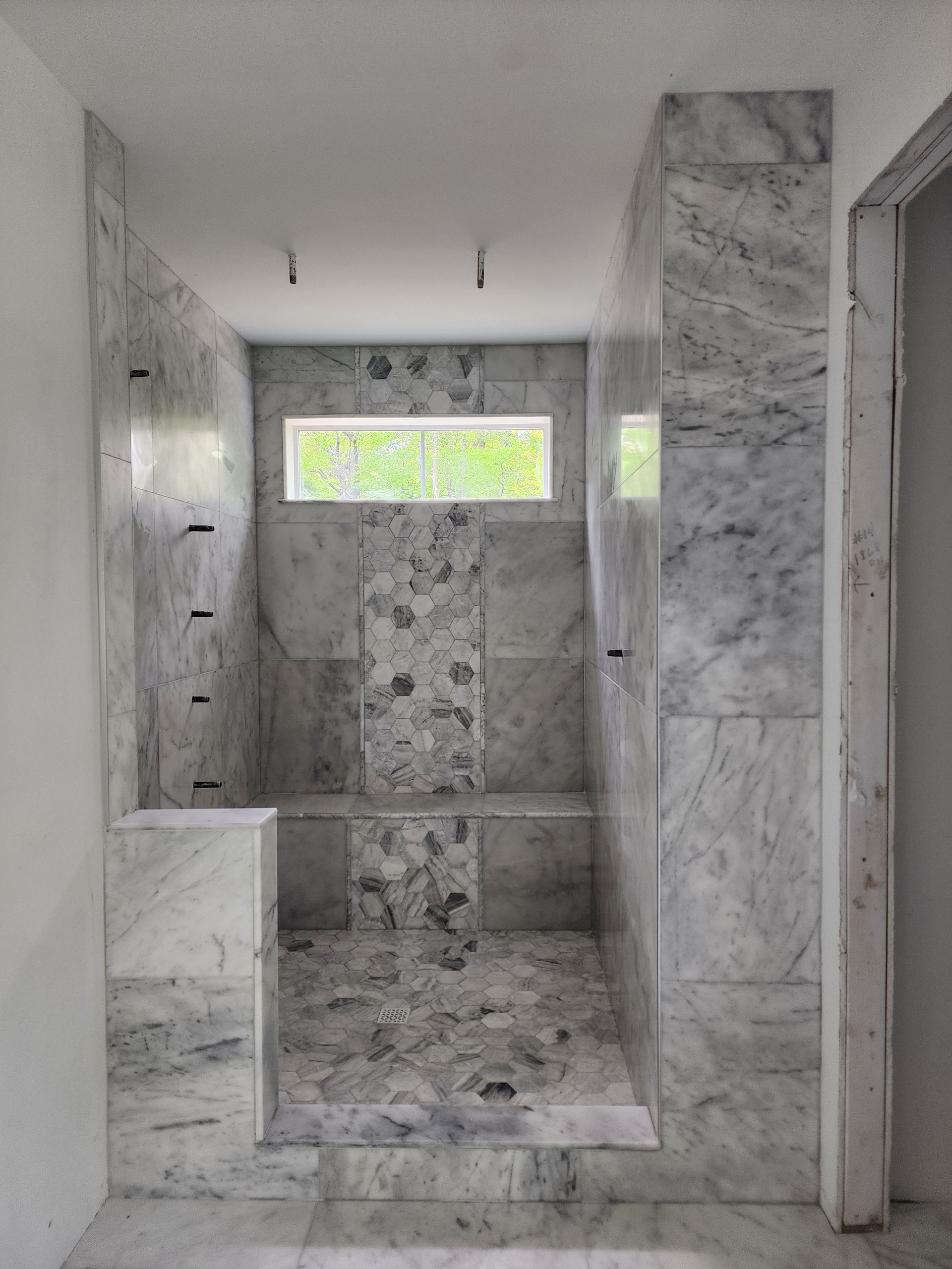 A walk-in shower under construction, featuring gray-and-white marble tiles, a built-in bench, and a small rectangular window.