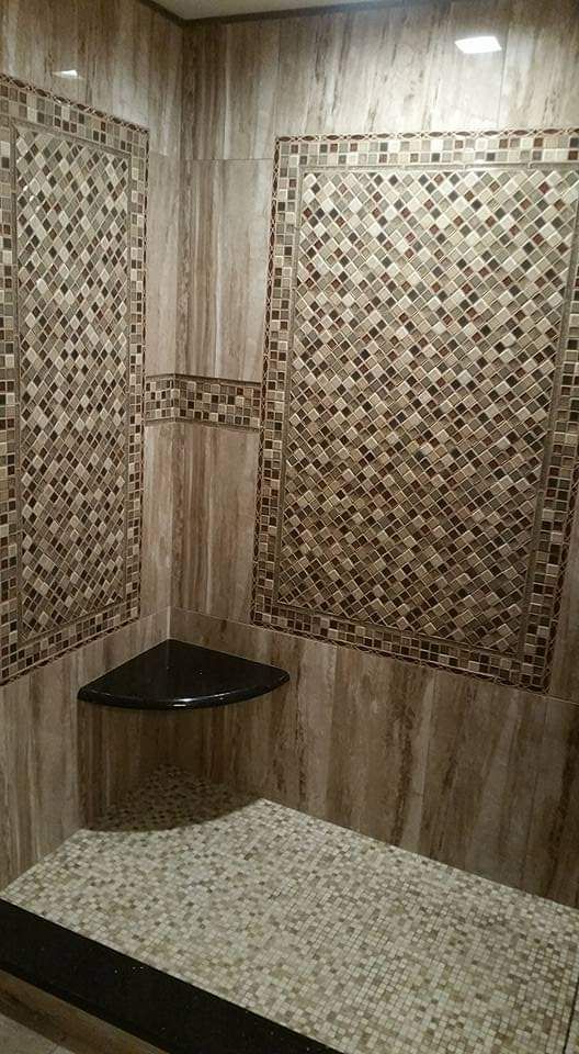 A shower stall with tan stone wall tiles, a mosaic tile inlay, a black corner shelf, and a light pebble floor.