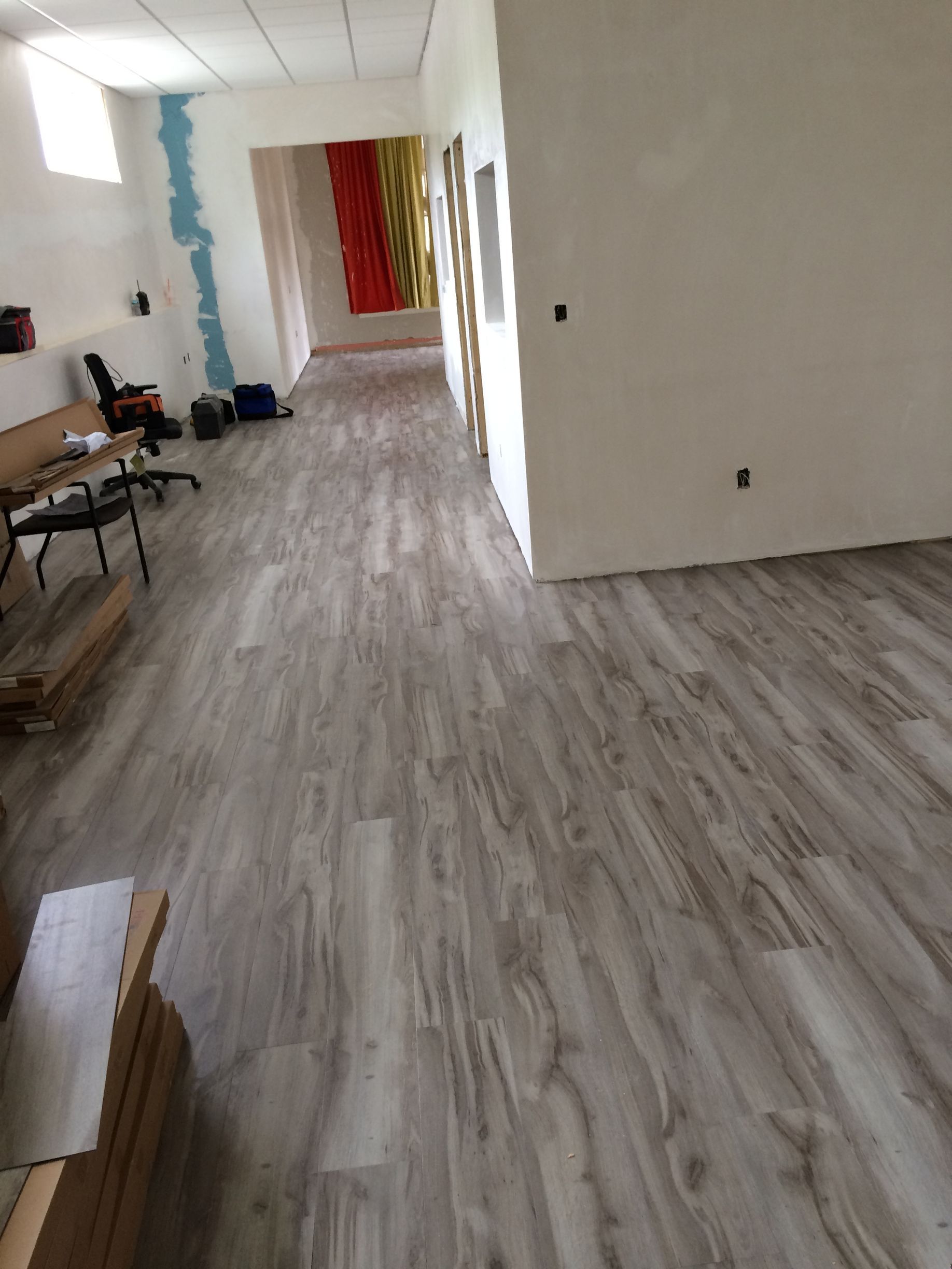 A long room with light wood-look flooring, white walls, and a desk area on the left. In the background, colorful curtains.