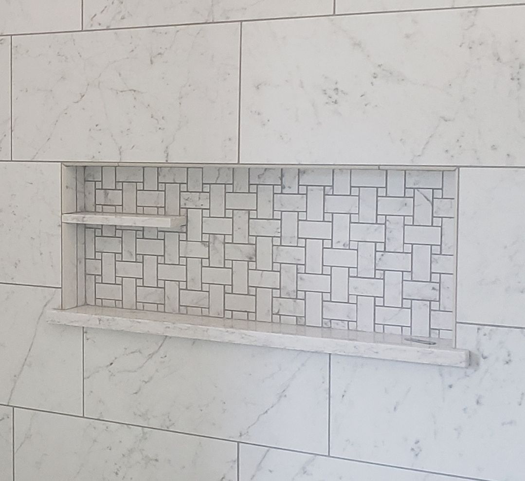 A recessed shower niche with white marble walls and a decorative basket-weave tile accent back panel with two shelves.