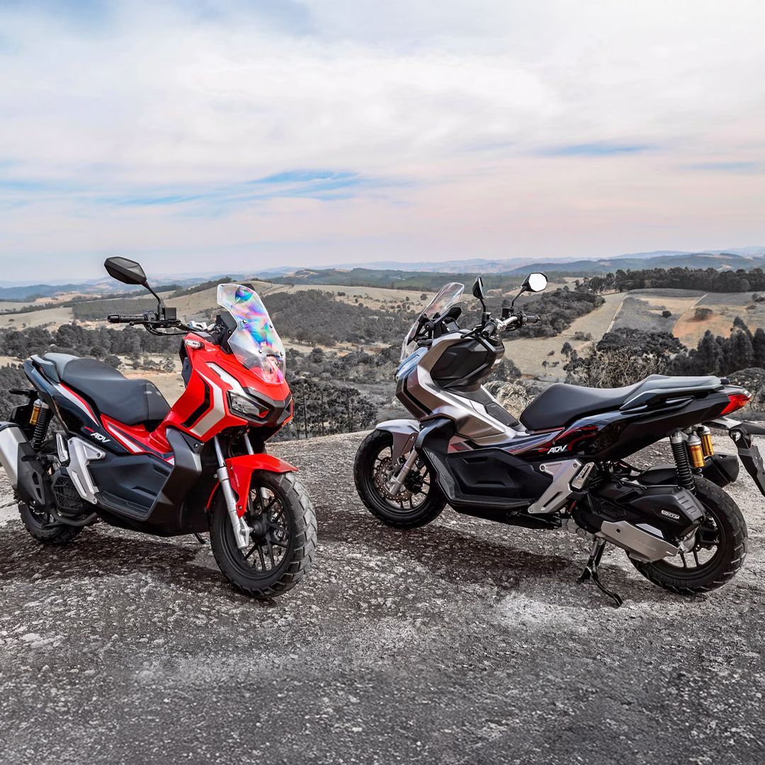 Duas scooters Honda ADV, vermelha e cinza, estacionadas no topo de uma colina com um cenário pitoresco.