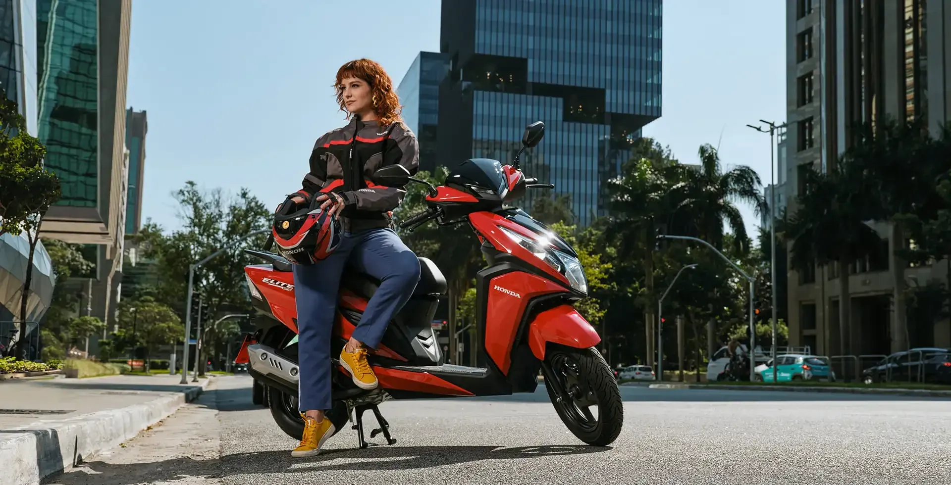 Mulher em uma scooter vermelha em uma cidade, vestindo jaqueta e capacete, sorrindo.