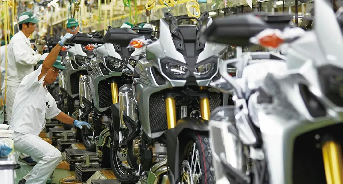 Linha de montagem de motocicletas: trabalhadores de jaleco branco montando motocicletas de aventura prateadas e pretas.