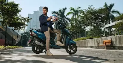 Homem em uma scooter azul, segurando capacete, fundo da cidade, dia ensolarado.
