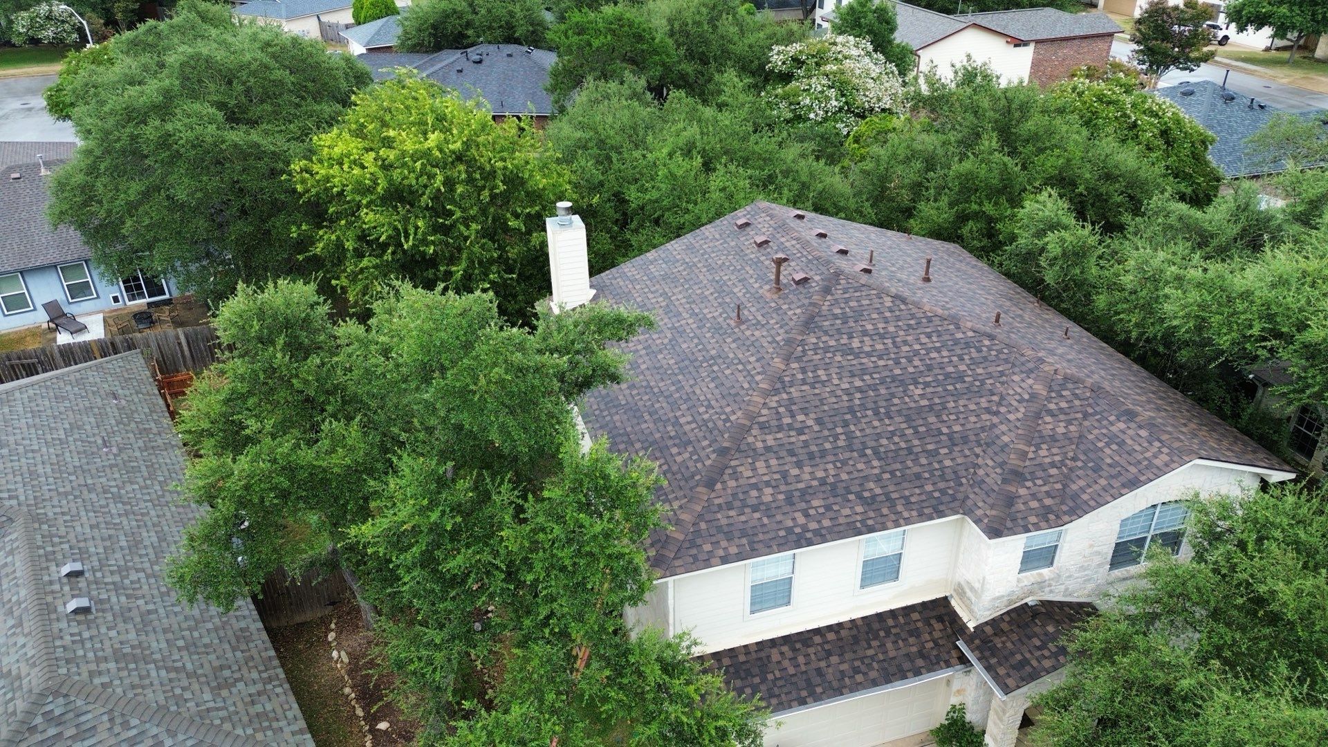 An aerial view of a tan residential house with a shingled roof, surrounded by lush green trees in a suburban neighborhood.