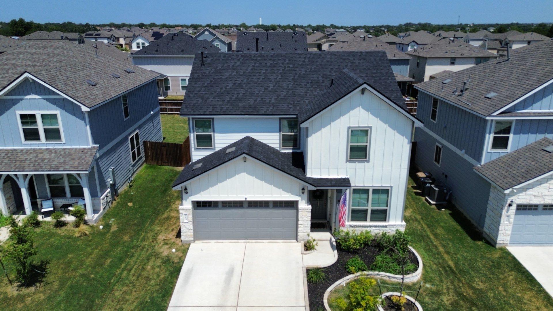 A two-story house with white vertical siding and a dark gray roof, surrounded by neighboring homes in a residential area.