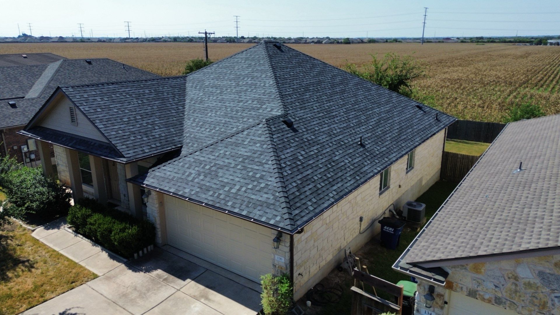 An aerial view of a single-story suburban house with a dark gray shingled roof, set beside a field of crops.