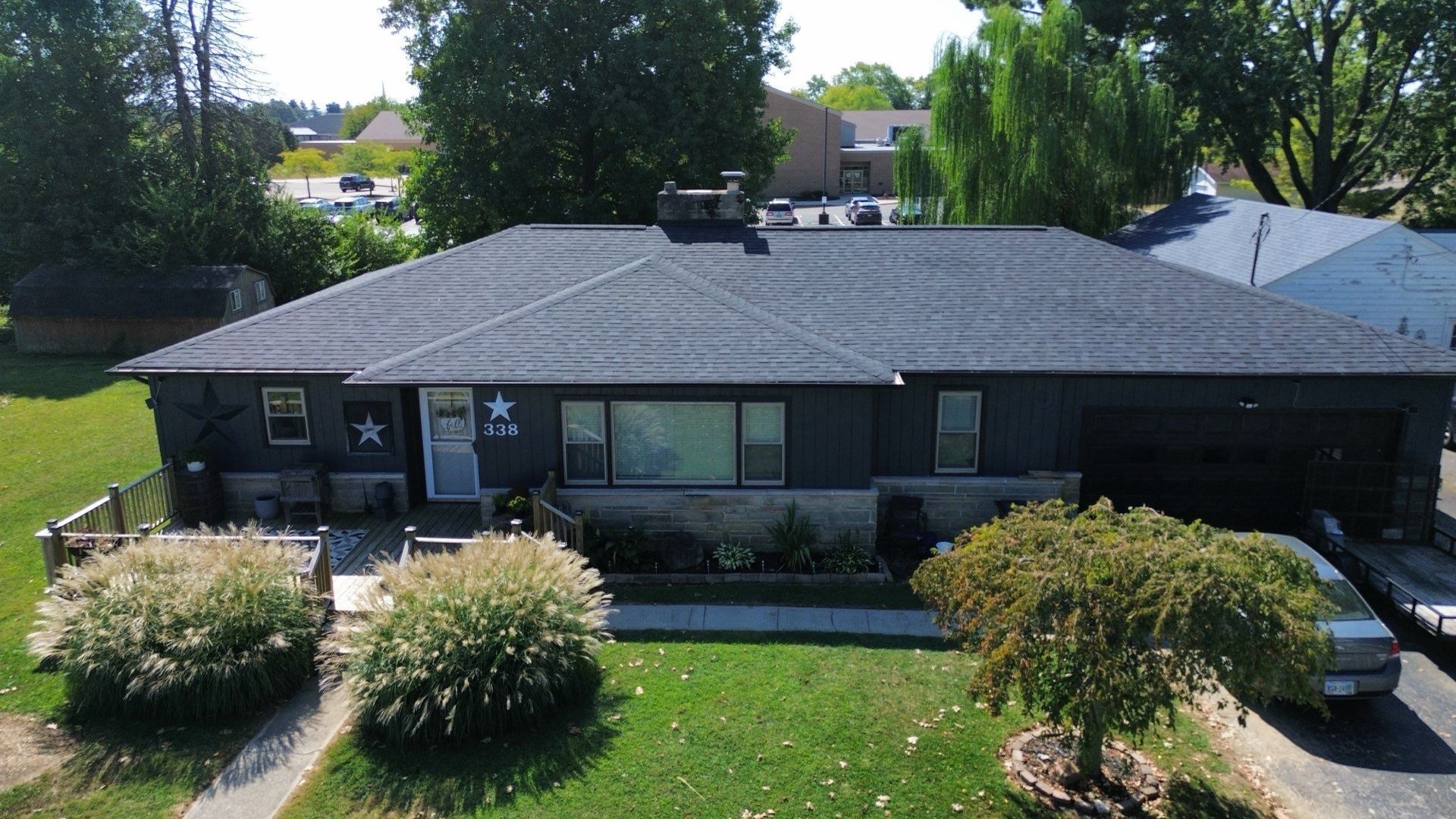 A dark gray, single-story house with a shingled roof, stone accents, two large shrubs in the front, and a driveway.