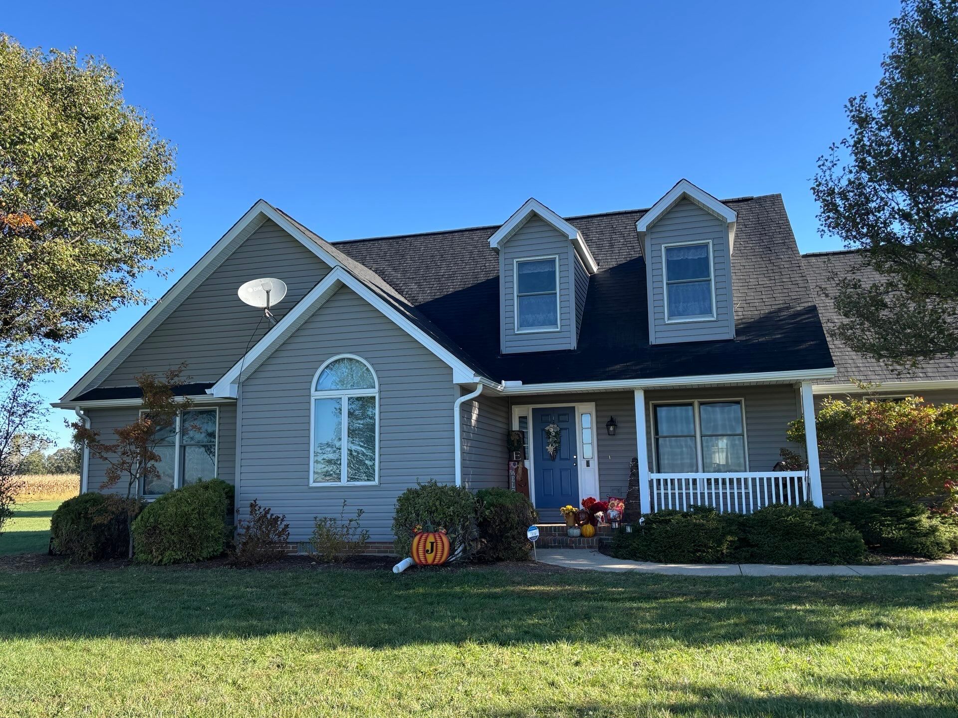 A two-story grey suburban house with a blue front door, two dormer windows, and a front porch, set in a grassy yard.
