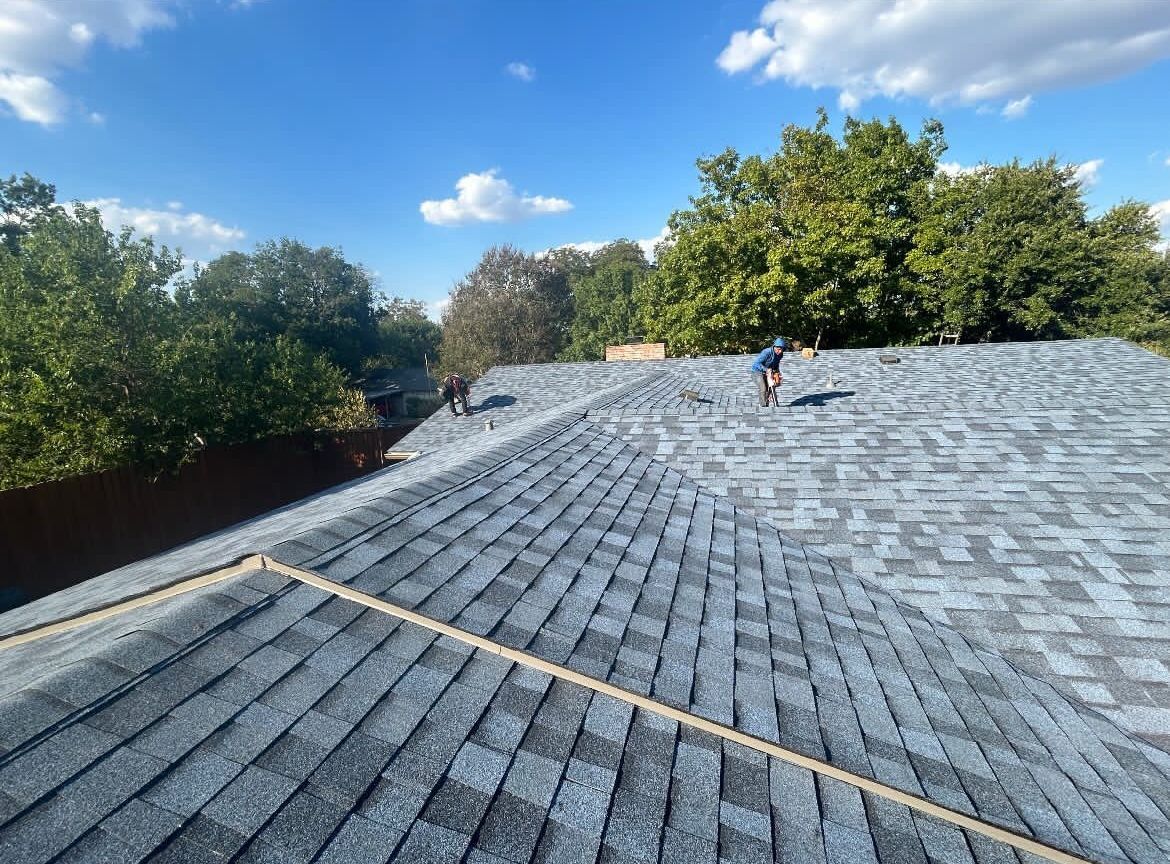 Workers install gray shingles on a suburban house roof under a sunny blue sky.