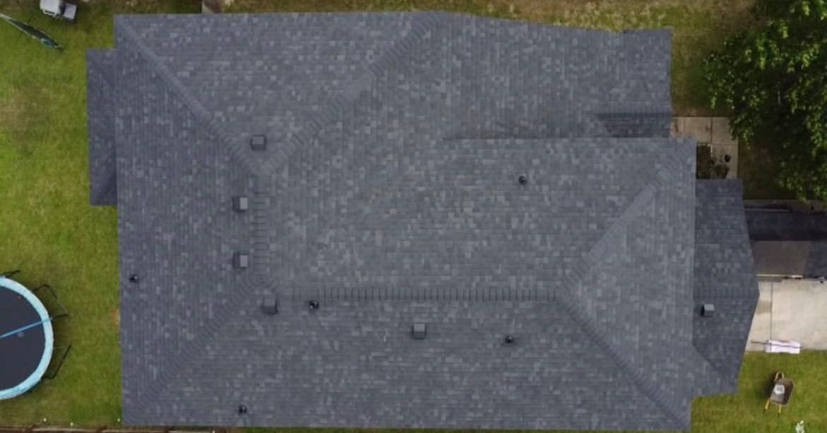 Top-down aerial view of a dark gray, shingled residential roof with several vents, set over a grassy lawn.