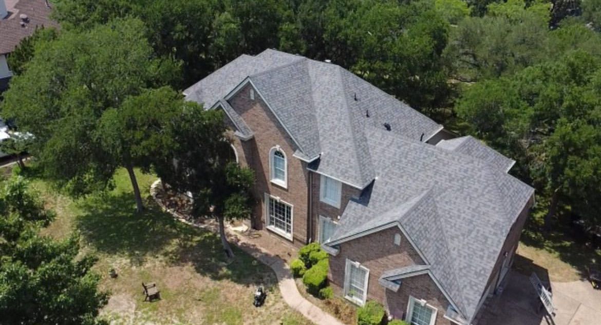 An aerial view of a two-story brick house with a gray shingled roof, surrounded by trees and a lawn.