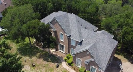 An aerial view of a two-story brick house with a gray shingled roof, surrounded by trees and a lawn.