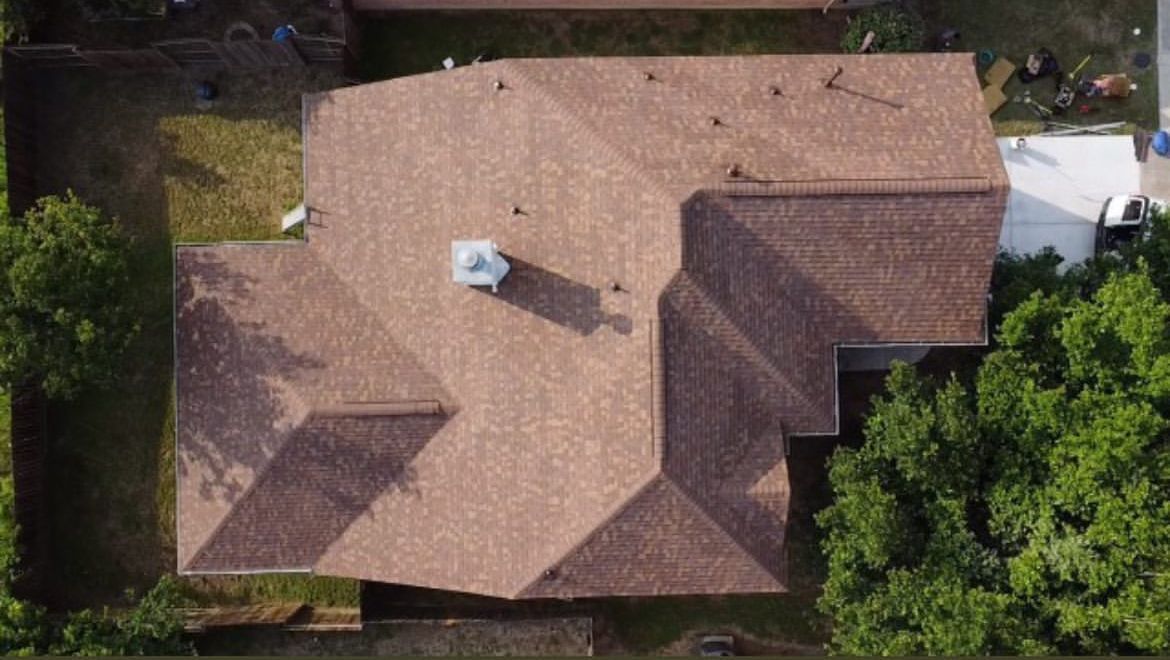 Aerial view of a brown shingled residential roof with a central chimney, surrounded by green trees and grass.