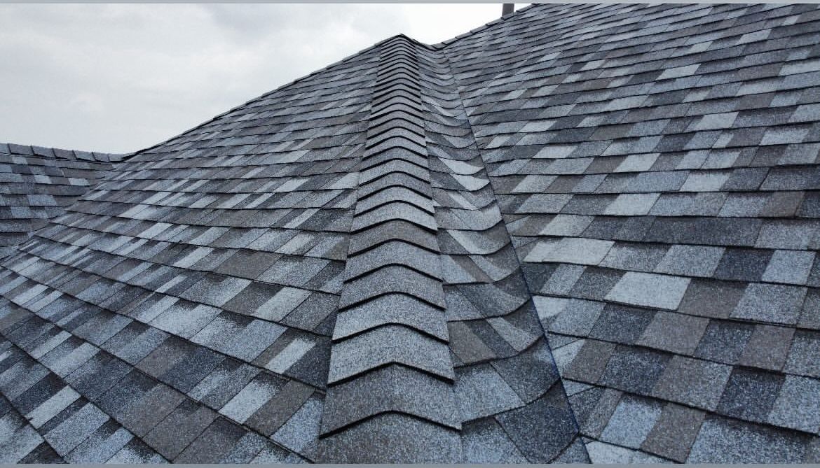 A close-up view of a shingled roof with a central ridge cap, showing textured, multi-tonal gray architectural shingles.