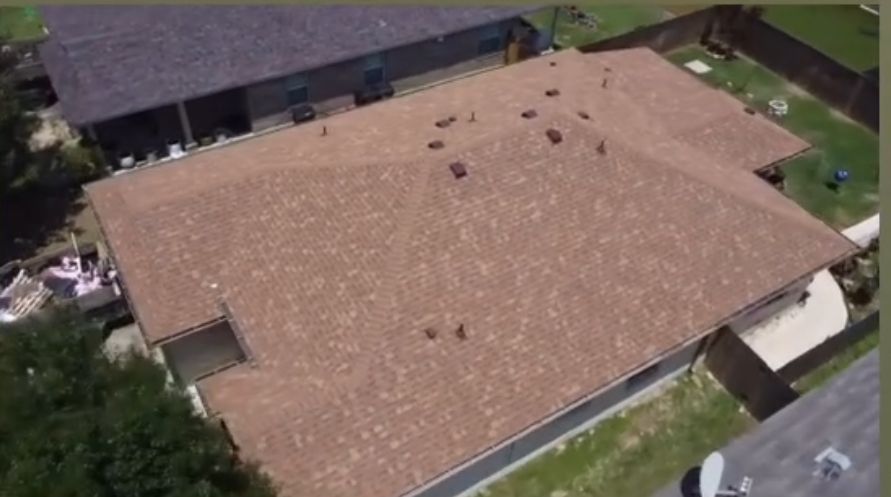 An aerial view of a tan, shingled residential roof with several vents, set between neighboring houses and yards.