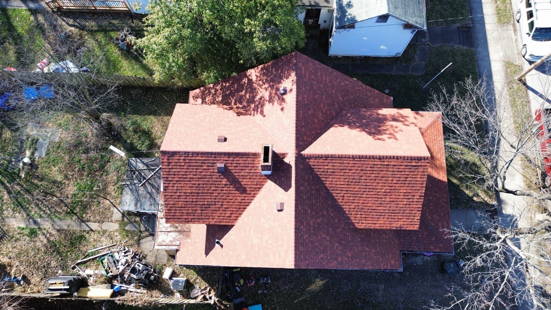 An aerial, top-down view of a house with a red shingled roof, showing multiple gables, a chimney, and surrounding trees.
