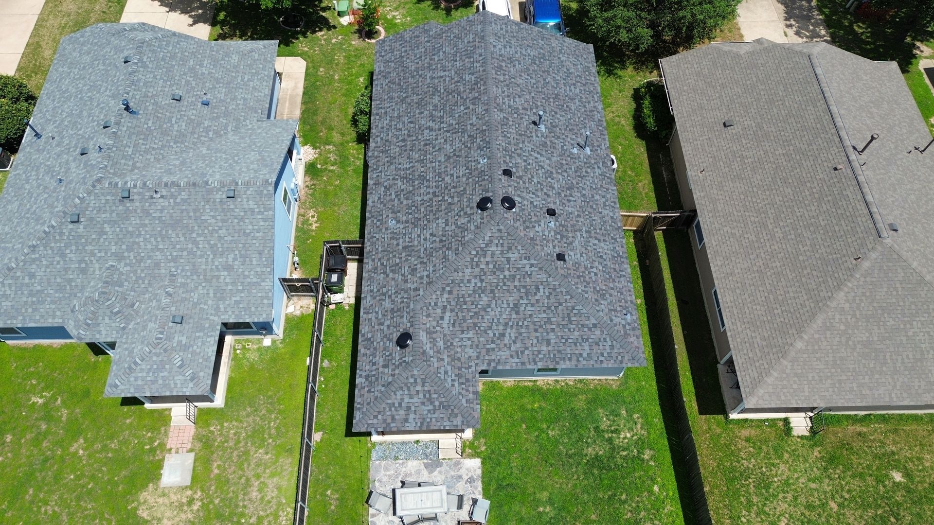 Aerial view of three suburban houses with grey shingled roofs, separated by green lawns and privacy fences.