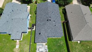 Aerial view of three suburban houses with grey shingled roofs, separated by green lawns and privacy fences.