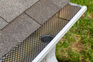 A white rain gutter covered with a black mesh guard sits beneath the edge of an asphalt shingle roof.