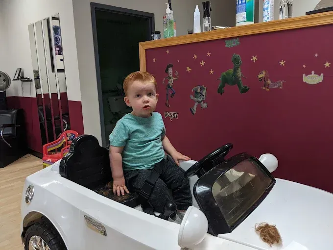 A little boy is sitting in a toy car in a barber shop.