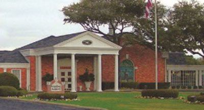 Brick building with white columns and a flag; green lawn with trimmed bushes.